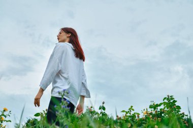 Beyaz tişörtlü genç kız bulutlu bir gökyüzünün altında çiçeklerle dolu bir tarlada dikiliyor, uzaklara bakıyor..