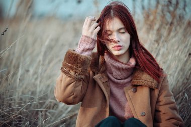 Kahverengi paltolu genç kız, açık havada uzun kuru çimenlerde oturuyor, gözleri kapalı, saçlarına hafifçe dokunuyor, sakin ve düşünceli bir hava veriyor..