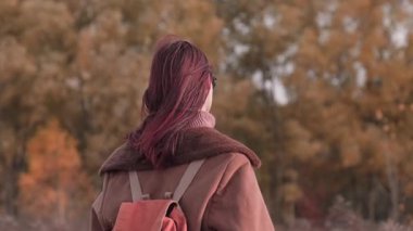 Genç kız kameraya arkası dönük duruyor, gerçek bir gülümsemeyle, güneş gözlüğü takıyor ve renkli bir sonbahar manzarasında sıcak kahverengi bir ceket giyiyor..