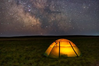 Baharda yeşil çimlerin üzerinde, içeriden parlak bir şekilde aydınlatılan sarı çadır Samanyolu 'nun arka planına ve şehirden uzak parlak yıldızlara karşı. Uzun pozlama astrofotoğrafçılık