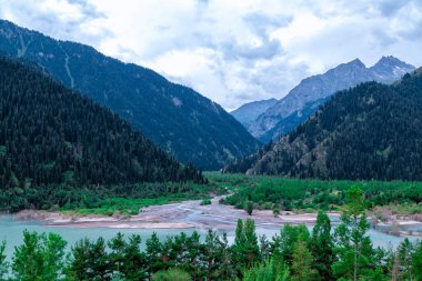 Issyk Gölü, Almaty, Kazakistan. Dağların arka planında mavi suyla dağ gölü ve akan bir nehrin ağzına tepeden bakan yeşil çimenli ağaçlar 1693 'teki bir çamur akışının sonuçlarıyla birlikte.