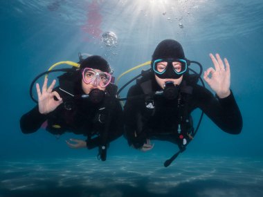 Suyun altında yüzen iki mutlu dalış dalgıcının yakın çekimi. Arkalarında parlak güneş ışınları parlayan tamam işaretini gösteriyorlar.
