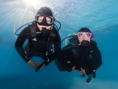 Suyun altında yüzen iki dalgıcın yakın çekimi. Arkalarında su yüzeyinde parlayan parlak güneş ışınlarıyla kameraya doğru yüzüyorlar.