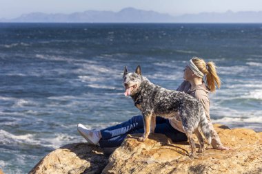 A woman sitting on a rock with an Australian Cattle Dog (Blue Heeler), looking at an ocean view outdoors, exploring together