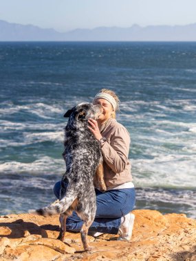 A woman with an Australian Cattle Dog (Blue Heeler) outdoors exploring together with the dog jumping up and kissing her