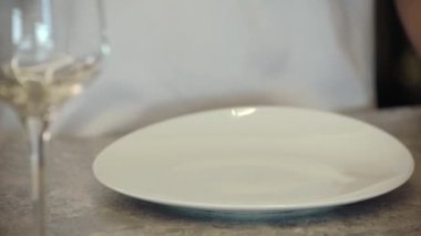 Bir adam masaya beyaz bir tabak ve şarap bardağı koyar..