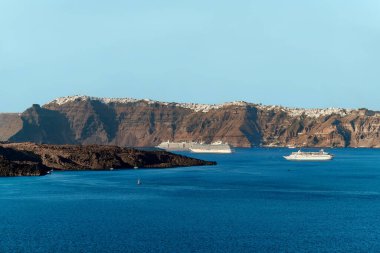 Nea Kameni volkanik kalderasının yolcu gemilerini ve arka planda Santorini adasının güneybatısından Imerovigli köyünü izleyin.