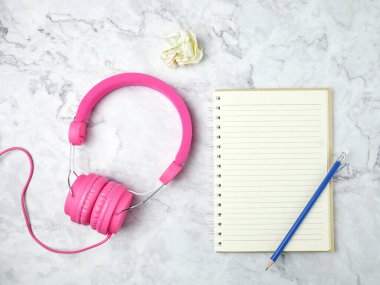 Pink headphones with a notebook and pencil on marble texture background.