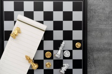 Chess board with chess pieces and notebook on stone texture background. Chess gold with silver battle. Top view.