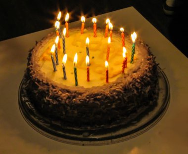 Güzel dekore edilmiş bir doğum günü pastası. Üstünde beyaz bir yüzey üzerinde yanan mumlar var. Pasta çikolata kaplı ve hindistan cevizi parçacıklarıyla süslenmiş, davetkar ve şenlikli bir ortam yaratıyor..