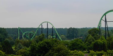 Hollanda, Biddinghuizen - 15 Temmuz 2014 - Birden fazla döngü ve tepesi olan yeşil bir lunapark treni, bir ağaç zeminine ve bulutlu bir gökyüzüne karşı kurulmuştur. Sahne, eğlence parklarının heyecanını yakalar..