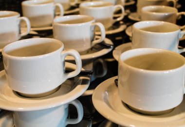 Sade ve zarif tasarımlarını sergilemek için çay tabakları üzerine dizilmiş birçok beyaz kahve fincanının yakın plan görüntüsü. Bardaklar düzgünce istiflenmiş, görsel olarak çekici bir desen oluşturuyor..