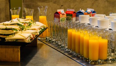 İçinde bir tabak sandviç, bir bardak portakal suyu ve bir kutu süt bulunan bir büfe. Sandviçler yeşilliklerle düzgünce düzenlenirken, portakal suyu berrak bardaklarda servis ediliyor, canlı ve davetkar bir vitrin oluşturuyor..