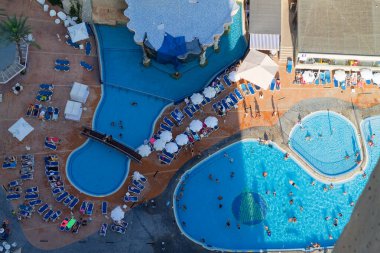 Birden fazla yüzme havuzu, güneş pansiyonları ve şemsiyelerin bulunduğu bir tatil köyü manzarası. İnsanlar suyun ve güneş banyosunun tadını çıkarıyor..