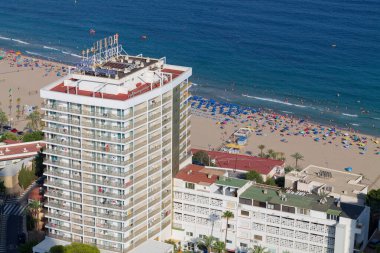 Renkli şemsiyeler ve güneşlenenlerle dolu kumlu bir sahile bakan bir sahil otelinin havadan görünüşü. Okyanus sakin ve mavidir. Uzakta birkaç tekne görünür..