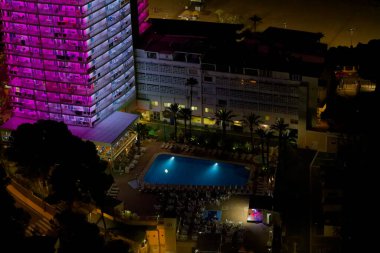 Geceleri, palmiye ağaçlarıyla çevrili parlak ışıklı bir havuz alanı olan bir otelin hava manzarası. Otelin renkli ışıklandırmalı modern bir tasarımı var, canlı bir atmosfer yaratıyor..