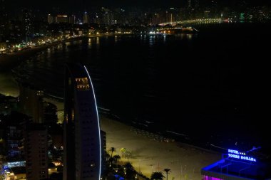 Sahil şeridi boyunca aydınlık binaların ve karanlık bir plajın yer aldığı bir kıyı şehrinin gece manzarası. Sahne, suyu yansıtan canlı şehir ışıklarını yakalıyor..