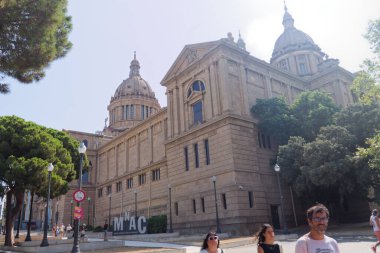 Kubbeleri ve geniş pencereleri olan büyük mimarisi olan tarihi bir bina. İnsanlar güneşli bir günde ağaçlar ve sokak işaretleriyle çevrili bir şekilde yürürler..