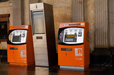 Renfe için iki turuncu bilet otomatı, bir İspanyol tren servisi, mermer duvarları ve sütunları olan bir istasyonda..