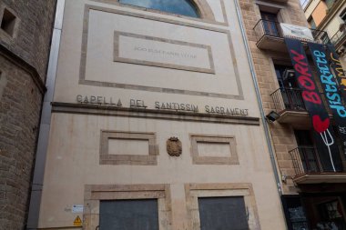 'CAPELLA DEL SANTISSIM SAGRAMENT' ve bir arma içeren basit bir tasarıma sahip tarihi bir şapel cephesi. Bina, modern mimariye yakın bir kentsel alanda yer almaktadır..