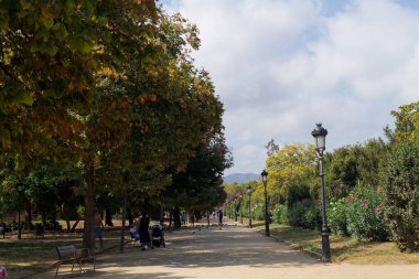 Ağaçlar, banklar ve sokak lambalarıyla kaplı sakin bir park yolu. İnsanlar, parçalı bulutlu bir gökyüzü altında doğal ortamın tadını çıkararak rahatça dolaşır..