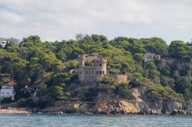 Kayalık bir yamaçtaki şatonun manzarası. Etrafı yemyeşil ağaçlarla çevrili. Önünde kumsalı olan sakin bir deniz manzaralı..