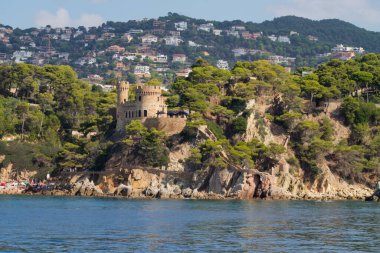Bereketli bir yeşillik ve sakin bir mavi denizle çevrili kayalık bir uçurumun üzerindeki bir şatonun manzarası. Arka planda, bir yamaçta konut binaları görülebilir..