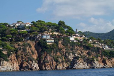Yeşillikler arasında yuva yapmış, kıyı uçurumunun manzarası. Manzara, parçalı bulutlu bir gökyüzünün altında kayalık oluşumlar ve sakin bir denizi barındırır..
