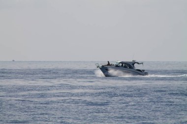 Pürüzsüz bir tekne bulutlu bir gökyüzünün altında sakin okyanus sularında yüzüyor. Tekne hareket ettikçe, huzurlu bir deniz manzarası sergiliyor..