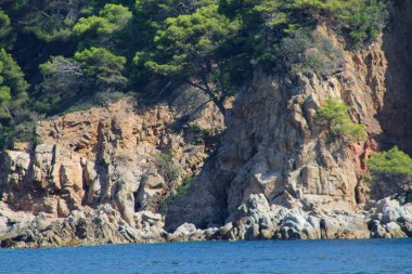 Kayalık uçurumlar ve yemyeşil yeşilliklerin olduğu manzaralı bir sahil manzarası. Su sakindir, doğal manzarayı yansıtır. Kayalıklarda ağaçlar büyüyor, manzaranın güzelliğini artırıyor..