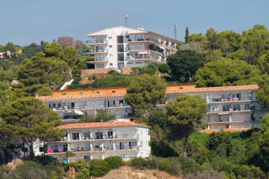 Yamaçtaki binaların manzarası yemyeşil ağaçlarla çevrili. Mimaride terracotta çatıları olan beyaz ve bej renkler yer alıyor ve sakin bir sahil ortamı sergiliyor..