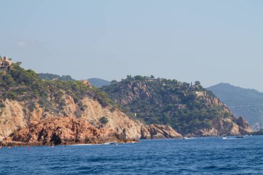 Kıyı şeridi boyunca kayalık kayalıklar ve yemyeşil yeşil manzaralı bir kıyı manzarası. Açık mavi deniz, suda yüzen birkaç tekneyle güneş ışığını yansıtır. Arka planda, yuvarlanan tepeler ve berrak bir gökyüzü manzarayı tamamlıyor..