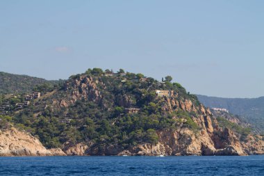 Berrak bir gökyüzünün altında sakin mavi bir denize bakan yemyeşil ve lüks evleri olan kayalık bir uçurumun manzarası..