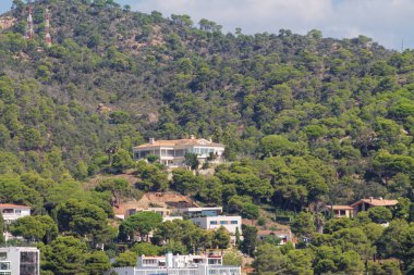 Yeşil ağaçlarla çevrili büyük beyaz bir evi olan bir tepenin manzarası. Manzarada, arka planda bir telekomünikasyon kulesi bulunan konut binaları ve doğal bitki örtüsü karışımı yer alıyor..
