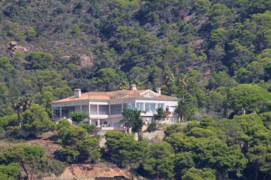 Yamaçta, yemyeşil ağaçlarla çevrili büyük, modern bir ev. Ev birden fazla balkona ve geniş pencerelere sahip ve manzaranın manzarasını sunuyor..