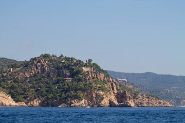 Bereketli yeşillik ve tepenin üzerine tünemiş beyaz bir ev ile kayalık bir sahil manzarası. Deniz sakin ve mavidir. Arka planda berrak bir gökyüzünün altında görünen uzak dağlar vardır..
