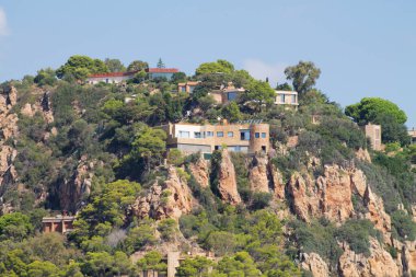 Bereketli yeşillik ve ağaçlarla çevrili kayalık bir yamaca tünemiş modern evlerin manzarası. Gökyüzü birkaç bulutla berrak, manzaranın güzelliğini arttırıyor..