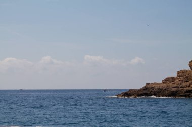 Kayalık bir kıyı şeridi, sakin mavi sular ve birkaç bulutlu berrak bir gökyüzü. Uzaktan küçük tekneler görülebilir, sakin atmosferi güçlendirir..