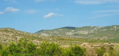 Berrak mavi gökyüzünün altındaki yuvarlanan tepelerin manzarası, ön planda yeşil alanlar ve arka planda kayalık arazi.