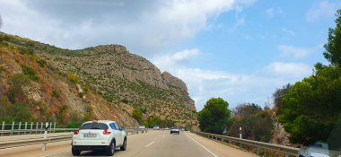 Dağlar ve yeşilliklerle çevrili, yolda giden araçlarla dolu manzaralı bir otoyol. Gökyüzü parçalı bulutlu, sakin bir atmosfer yaratıyor..