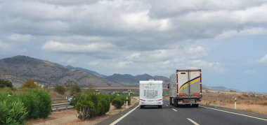 Beyaz bir karavan ve büyük bir kamyonun yan yana seyahat ettiği manzaralı bir otoyol manzarası. Arkaplanda dağlar ve yol kenarında yeşillik olan bulutlu bir gökyüzü var..