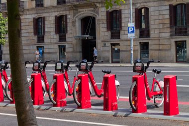 Şehir caddesinde bir bisiklet paylaşım istasyonuna park edilmiş kırmızı bisikletlerden oluşan bir sıra. Arka planda geniş pencereli ve yaya geçitli bir bina yer alıyor. Sahne aydınlık ve güneşli, ağaçlar caddeyi kaplıyor..