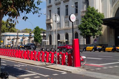 Şehir caddesi boyunca kırmızı bisiklet paylaşım istasyonları, park edilmiş bisikletleri ve şehir mimarisinin arka planı var. Arka planda sarı taksiler görünüyor..