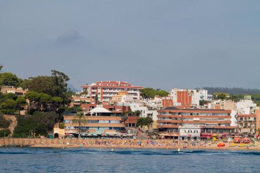 Güneş banyosu, renkli şemsiyeler ve plaj aktiviteleriyle dolu kumlu bir sahil manzarası. Arka planda, yeşilliklerle çevrili oteller ve restoranlar da dahil olmak üzere çeşitli binalar var..