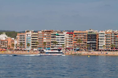 Sahilde manzaralı, ön planda bir tekne var. Sahil şeridi renkli binalar ve palmiye ağaçları ile kaplı ve insanlar kumlu sahilde güneşin tadını çıkarıyorlar..