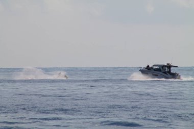 Sakin okyanus sularında yol alan bir tekne ve yanında jet ski ile uçan bir kişi. Sahne bulutlu bir gökyüzünün altında sakin bir deniz ortamı yakalıyor..