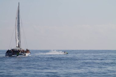Büyük beyaz yelkenli bir yelkenli, birkaç kişiyle birlikte sakin mavi sularda seyrediyor. Ön planda, bir jet ski geride bir sprey bırakarak suyun üzerinde hızla ilerliyor. Gökyüzü açık ve parlak.