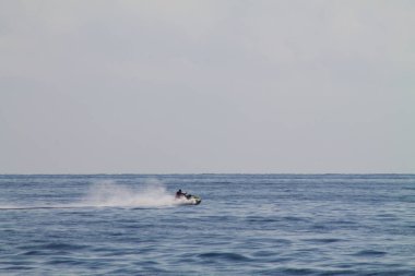 Sakin bir okyanusta jet ski süren bir insan, bir su püskürtüsü yaratıyor. Ufuk arka planda görünür, yukarıda açık bir gökyüzü var.