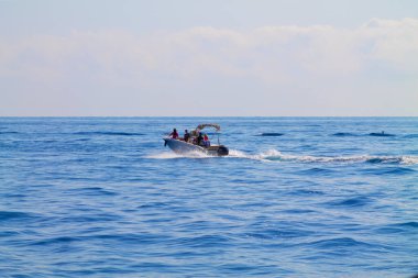 Birkaç kişiyle birlikte küçük bir tekne, sakin okyanus sularında yol alıyor ve arkasında beyaz köpükten bir iz bırakıyor. Gökyüzündeki birkaç bulutla ufuk açık..