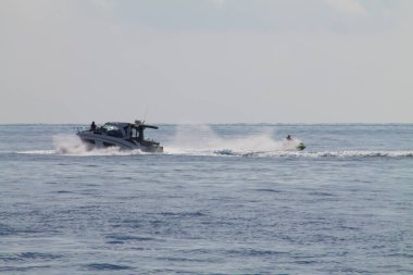 Bir motorlu tekne, yanında jet ski ile sakin sularda yüzüyor. Su sıçramaları ve dalgalar yaratıyor. Sahne açık bir gökyüzü ve uzak bir ufuk üzerine kurulmuştur..